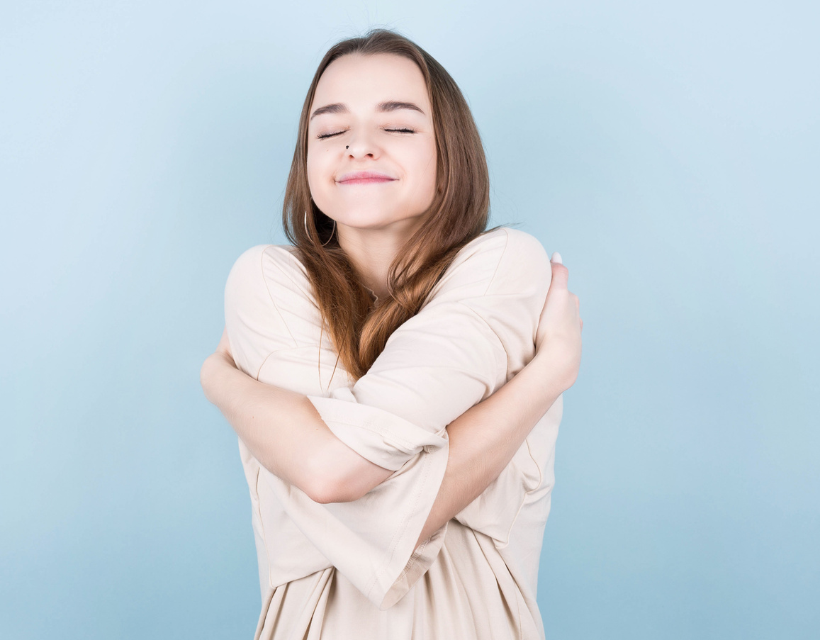 Young beautiful brunette woman wearing casual tank top, standing against blue background Hugging herself happy and positive, smiling confidently. Self-love and self-care